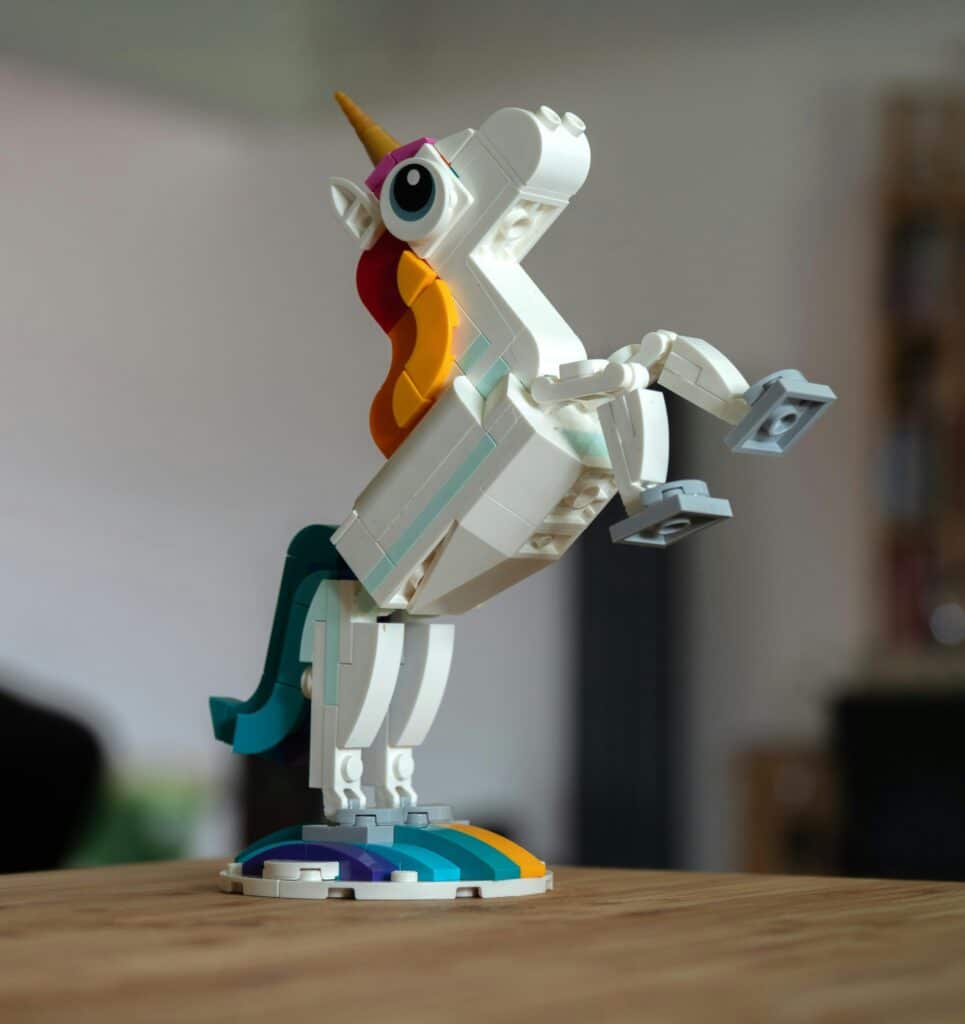 Een lego eenhoorn figuur staat op een houten tafel met opgetilde poten en een kleurrijke regenboogvoet.