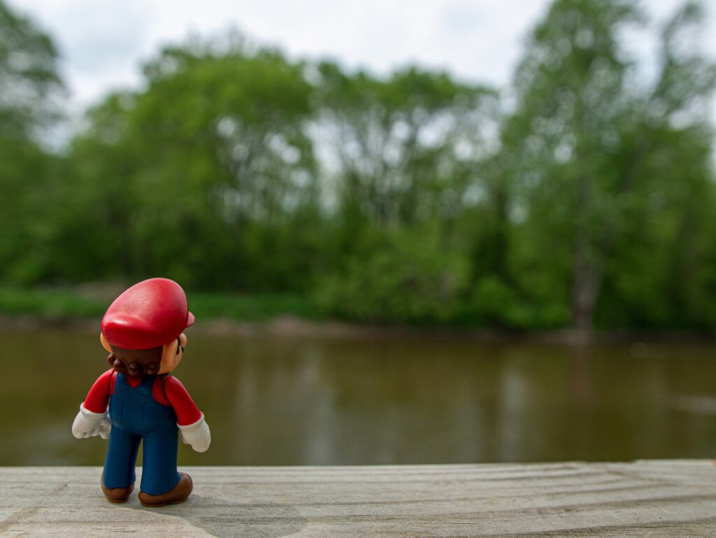 Een Mario speelgoedfiguur staat op een houten rand en kijkt uit over rustig water met groen bos op de achtergrond.